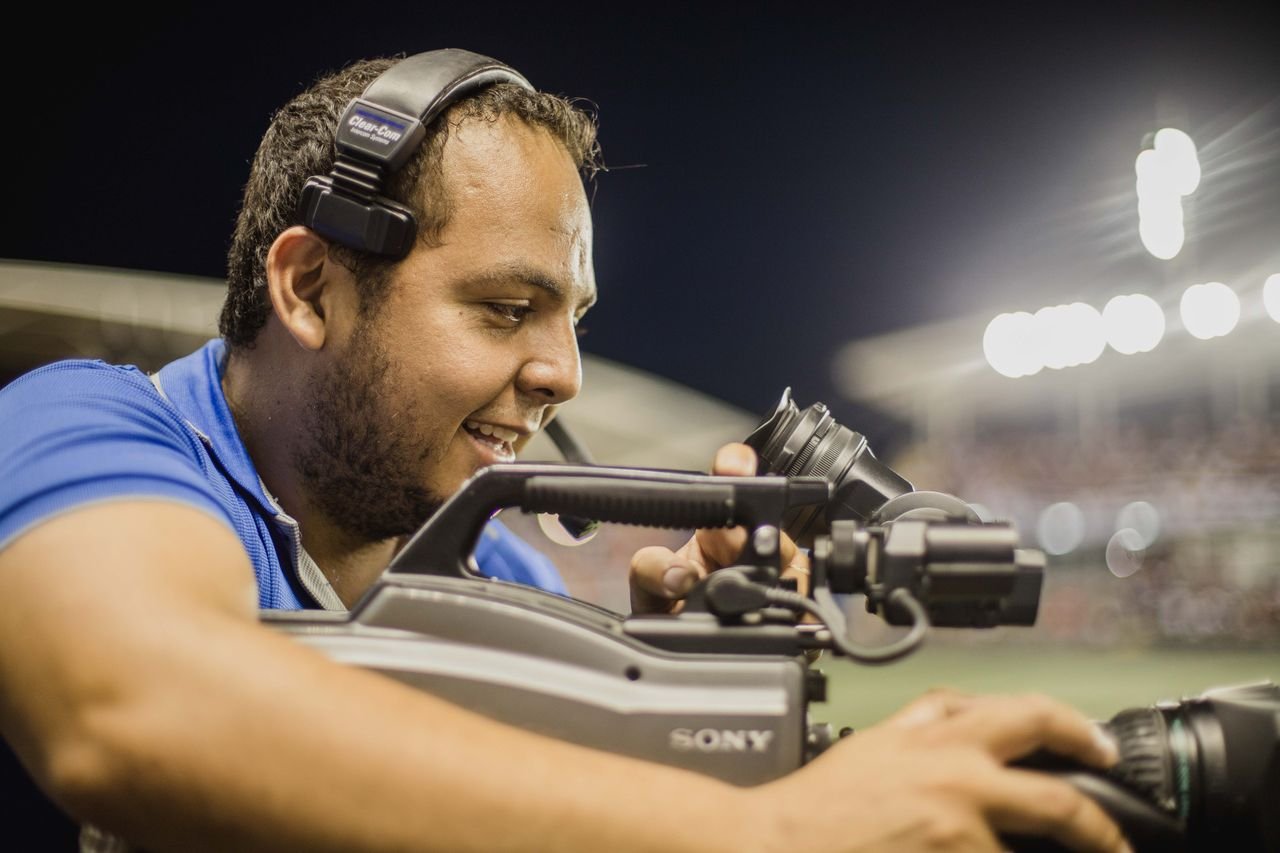 A cameraman with a headset operates a video camera outdoors at night, focusing on capturing footage.