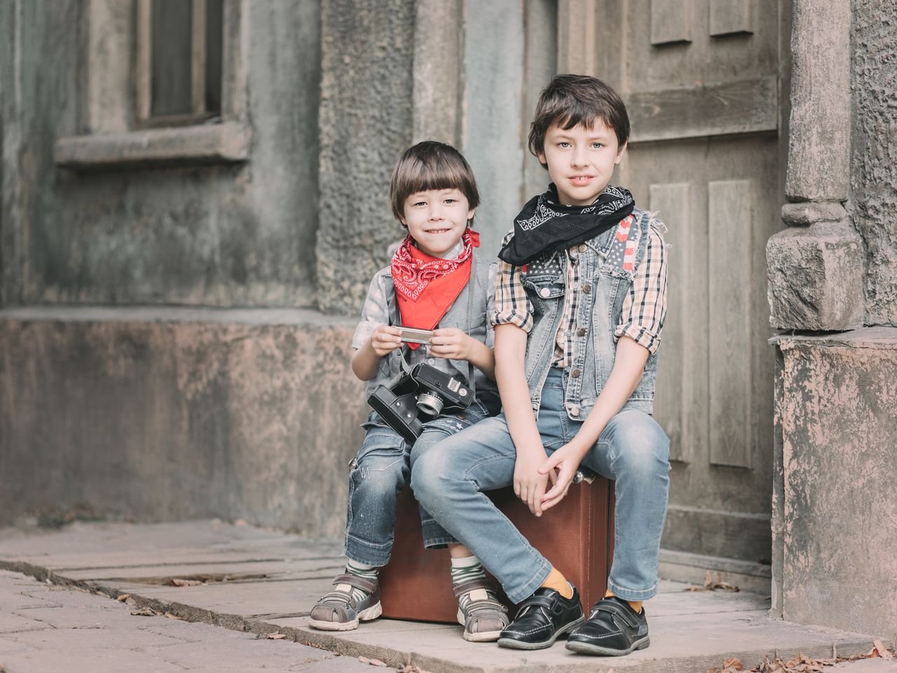 Two children sitting on a suitcase, showcasing vintage style and joy with a camera outdoors.